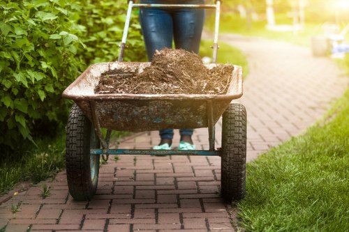 A person wearing blue jeans and black shoes is standing behind a garden wheelbarrow filled with dark soil, positioned on a paved pathway with red bricks. The wheelbarrow has a metal frame with black rubber tires and wooden handles. The pathway is bordered on the left by lush green bushes and on the right by a well-maintained grassy area. In the background, there is a bright, sunlit yard with additional plants and garden elements, suggesting an outdoor gardening or clearing activity in a residential area. This scene is associated with garden clearance services offered by Garden Clearance Kentish Town, focusing on debris removal and garden tidy-up tasks in Kentish Town area.