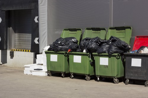 Six large wheelie bins are lined up on a paved surface outside a commercial building, with the first three being green and the subsequent one black. The green bins are filled with black rubbish bags, some of which are tied at the top, and are stacked to overflowing, with one bag resting on the lid of a green bin. The black bin on the right appears to contain some mixed waste without bags. Behind the bins, there are grey roller shutter doors and a section of the building’s exterior wall. To the left, a small stack of white bricks or concrete blocks is visible on the ground. The scene suggests a waste collection or rubbish removal service, typical of local clearance operations in Kentish Town. The overall environment indicates an industrial or commercial yard area, consistent with services provided by companies like Garden Clearance Kentish Town in the local postcode area.