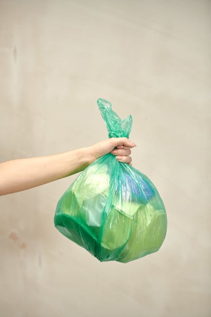 A person's arm and hand are visible, holding a tightly knotted green plastic rubbish bag filled with waste. The bag appears to contain mixed refuse, with some parts of the plastic surface showing tran