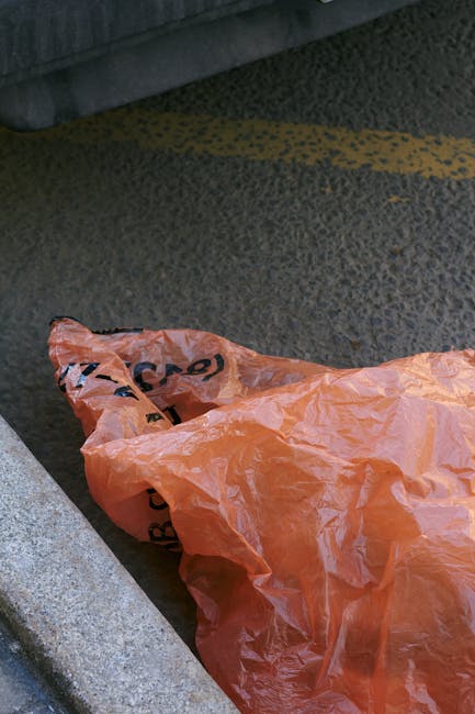 A person's arm and hand are visible, holding a tightly knotted green plastic rubbish bag filled with waste. The bag appears to contain mixed refuse, with some parts of the plastic surface showing tran